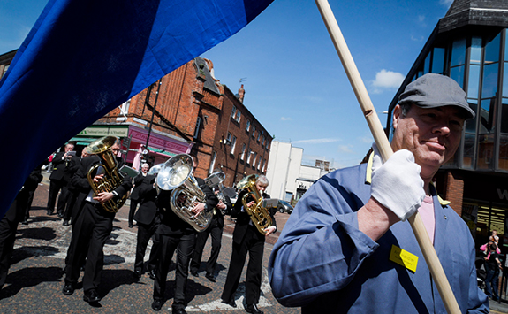 Street view of procession. In foreground a man hold a flag, with musicians in background